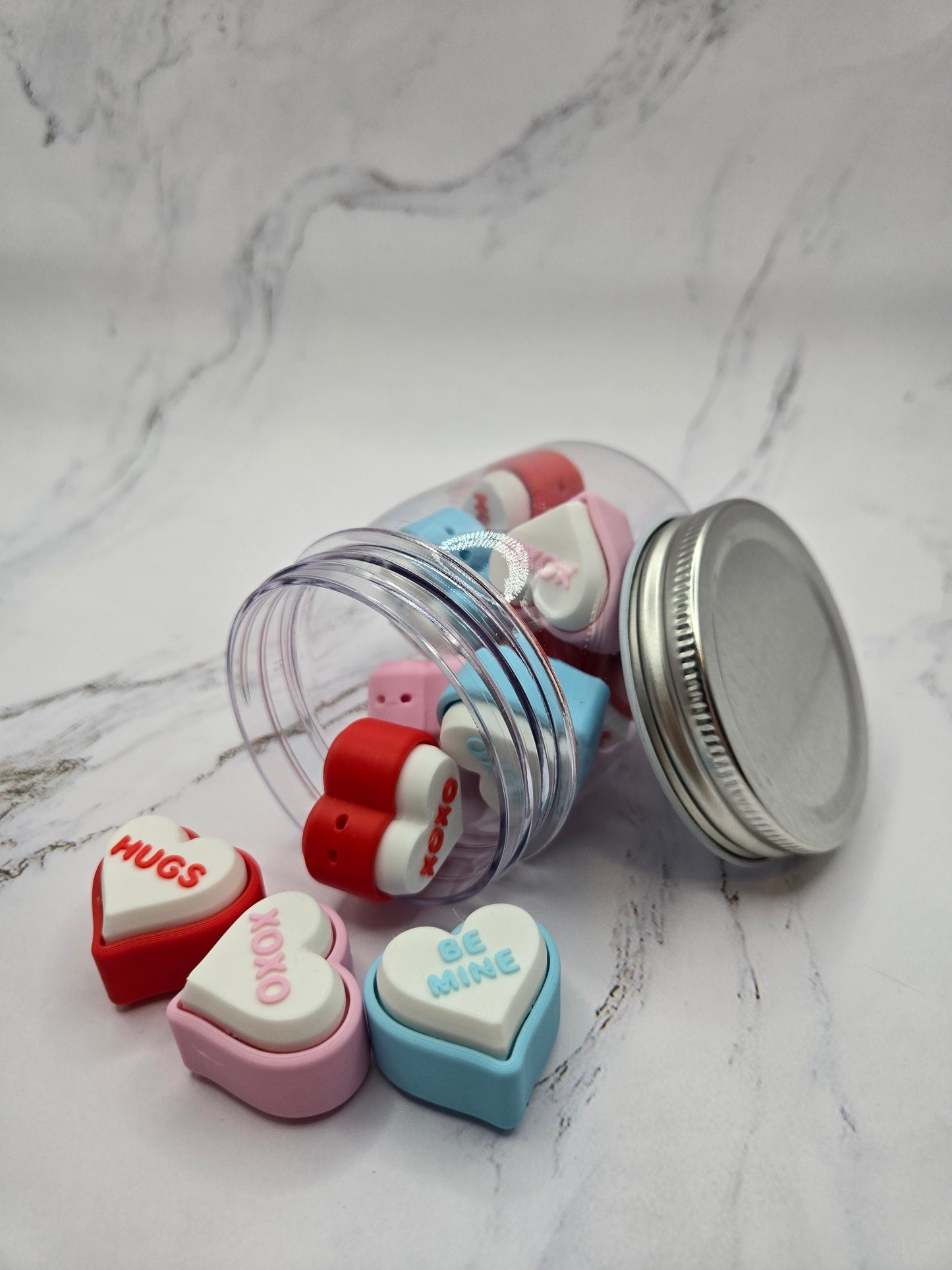 Close-up of 3D printed conversation heart fidget clickers spilling from jar showing colorful hearts with messages Hugs XOXO and Be Mine in red pink blue and white Valentine's Day clicky sensory fidget toy gift