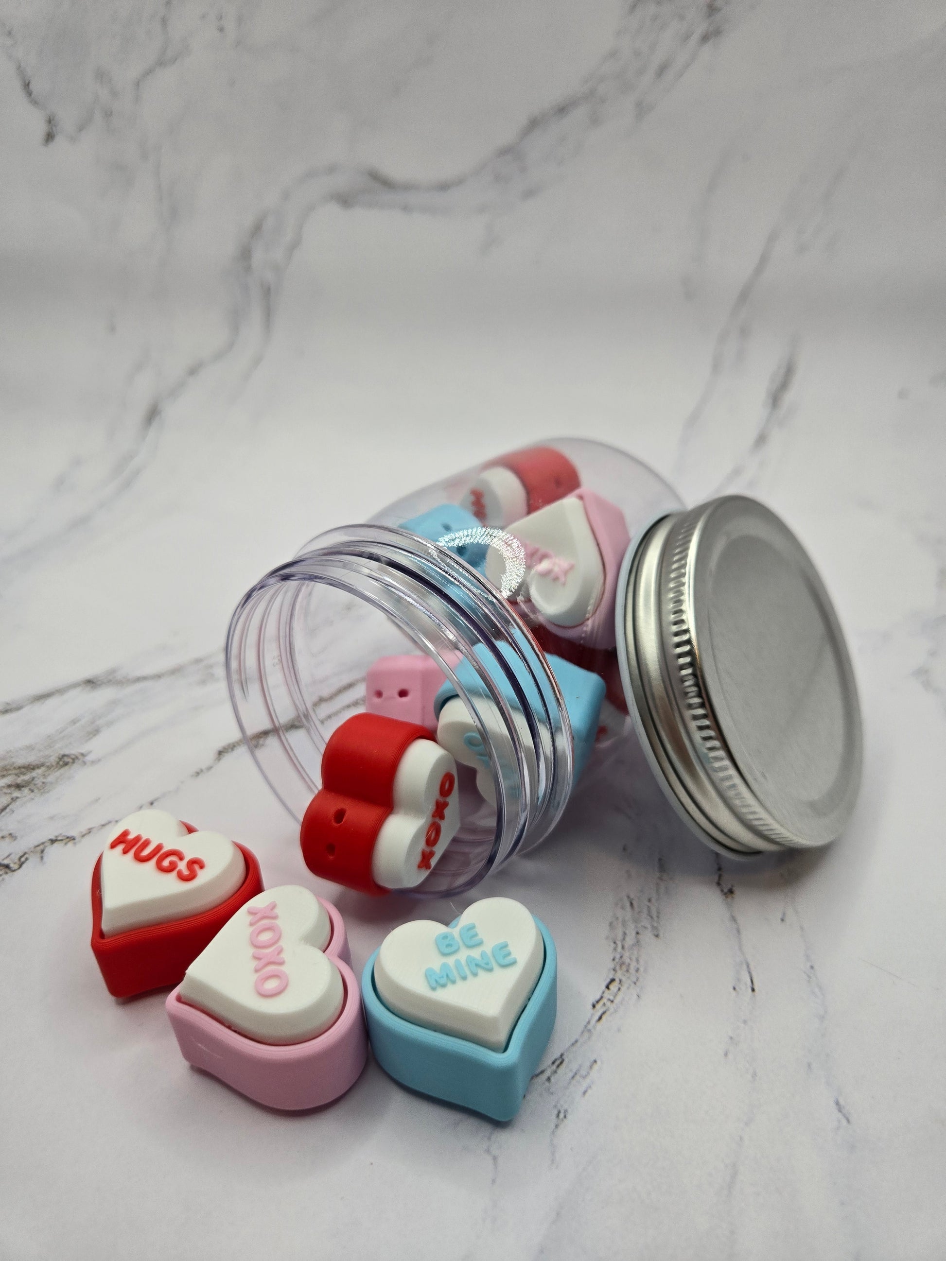 Close-up of 3D printed conversation heart fidget clickers spilling from jar showing colorful hearts with messages Hugs XOXO and Be Mine in red pink blue and white Valentine's Day clicky sensory fidget toy gift