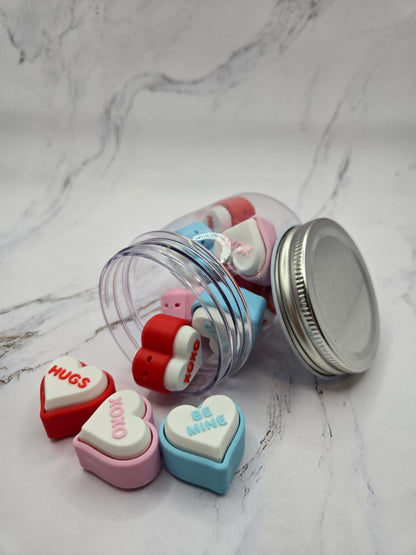 Close-up of 3D printed conversation heart fidget clickers spilling from jar showing colorful hearts with messages Hugs XOXO and Be Mine in red pink blue and white Valentine's Day clicky sensory fidget toy gift