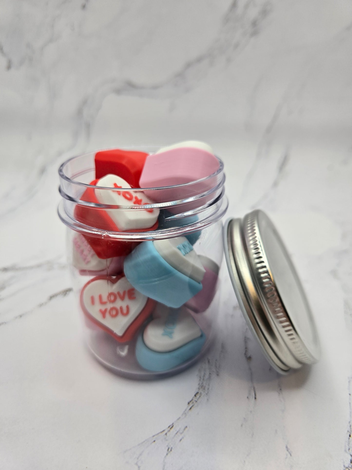 Open jar of colorful 3D printed conversation heart fidget clickers overhead view showing red pink blue and white hearts with messages like I Love You and Kiss Me Valentine's Day sensory toy gift